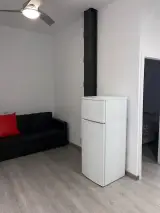 Interior apartment living room with black sofa and red cushion, white fridge and dark beam by the open bedroom door